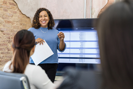 The mid adult female manager smiles as she uses a large screen to present spreadsheets to unrecognizable employees.