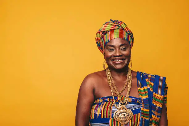 Smiling Ghanaian woman wearing a traditional attire against a yellow background. Happy black woman dressed in colourful Kente cloth and golden jewellery. Mature elegant woman embracing her rich culture.