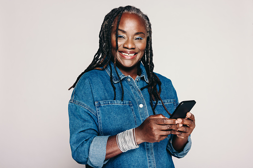 Mature woman smiling at the camera while holding a smartphone and standing against a grey background. Happy woman with dreadlocks wearing a denim jacket and make-up in a studio.