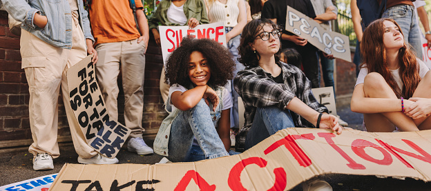 Cheerful teenage girl smiling at the camera while sitting with a group of youth peace activists. Multiethnic young people displaying posters and banners while protesting against war and violence.