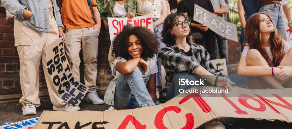 Cheerful teenage girl sitting with a group of youth peace activists Cheerful teenage girl smiling at the camera while sitting with a group of youth peace activists. Multiethnic young people displaying posters and banners while protesting against war and violence. Generation Z Stock Photo