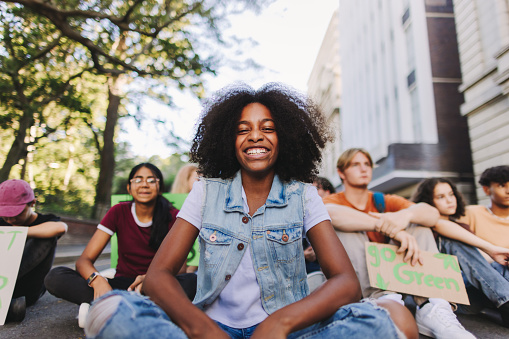 Happy black girl smiling at the camera while sitting with a group of demonstrators at a climate change protest. Multicultural youth activists joining the global climate strike.