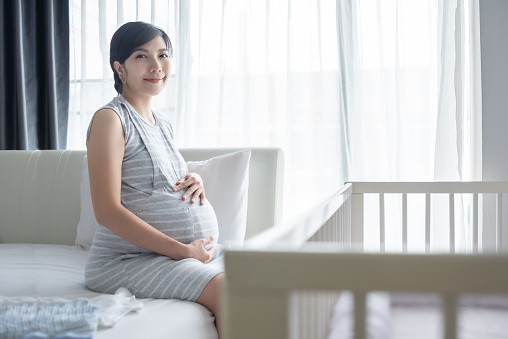 Portrait of Happy Smiling Asian Chinese or Korean Pregnant woman sitting on bed holding and touching her belly at bedroom.
