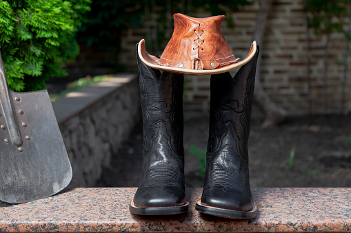 Attributes of american farmer. Black men cowboy-style leather boots stand on a granite walkway. On top of a brown leather hat and next to a shovel