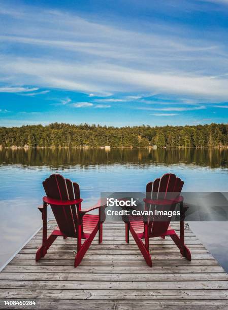 Two Red Adirondack Chairs On The End Of A Dock Overlooking A Lake Stock Photo - Download Image Now