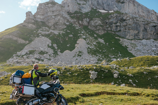 A motorcycle rider is standing in the mountain looking at the mountain top above him and enjoying the beautiful nature