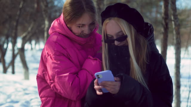 Two trendy teenage smiling girls in pink and black jackets look at photo on smartphone together in winter snowy forest on sunny day. Close-up.