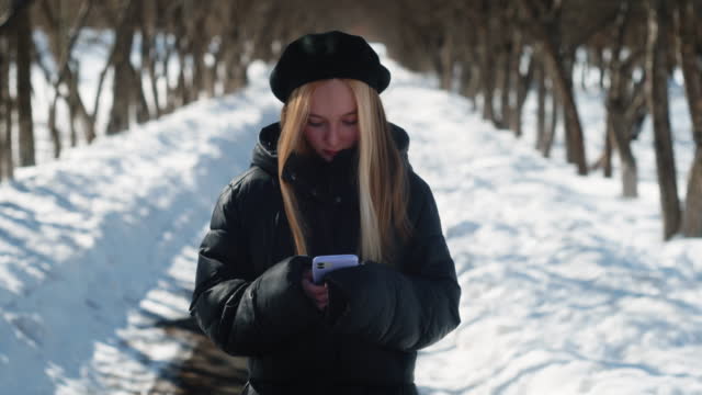 Young long haired blonde in beret and black jacket uses mobile phone on winter walk. Beautiful girl looks smartphone on snowy park street