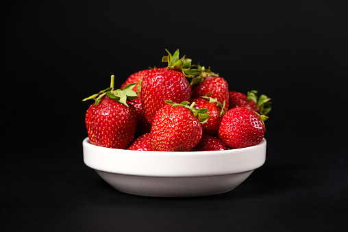 Red fresh strawberries in a white plate on a black background. Summer seasonal berries, close-up.