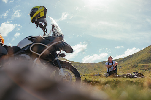 A seasoned Caucasian motorcycle rider is sitting on the grass next to his bike resting after a day of riding in beautiful landscape.