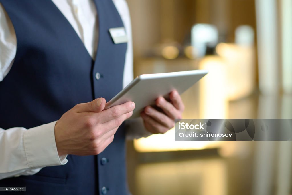 Digital tablet in hands of young male receptionist of luxurious modern hotel Digital tablet in hands of young male receptionist of luxurious modern hotel standing in lounge by entrance and meeting new guests Hotel Stock Photo