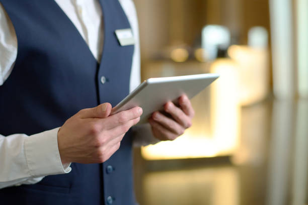 Digital tablet in hands of young male receptionist of luxurious modern hotel Digital tablet in hands of young male receptionist of luxurious modern hotel standing in lounge by entrance and meeting new guests hotel stock pictures, royalty-free photos & images