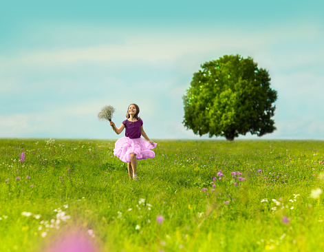 A happy girl runs across the summer field with a bouquet in her hands. There is a huge tree in the background. Girl wearing a pink skirt