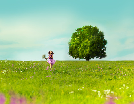 A happy girl runs across the summer field with a bouquet in her hands. There is a huge tree in the background. Girl wearing a pink skirt