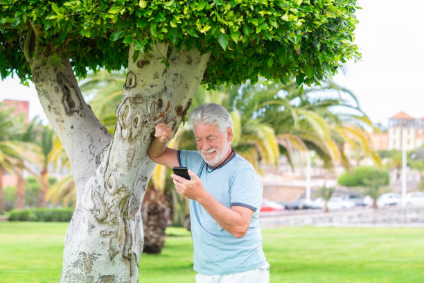 istock Smiling bearded elderly man leaning against a tree trunk in the park holding mobile phone, handsome elderly male talking on cellphone under the shade of the plant 1408470730