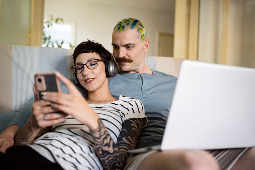 Caucasian man with short and crazy dyed hair, working on laptop, while his girlfriend leaning on him, listening to music and surfing the net on mobile phone
