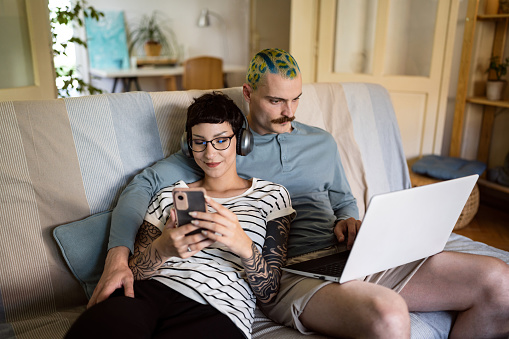Caucasian man with short and crazy dyed hair, working on laptop, while his girlfriend leaning on him, listen to music and surfing the net on mobile phone