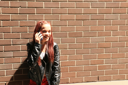 A Caucasian woman talking on a mobile phone. She is wearing a black leather jacket, and a necklace. She is in front of a brick wall.