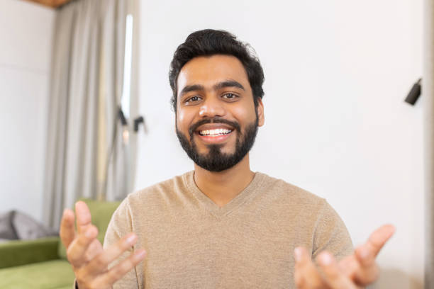 Portrait of happy young Indian guy sitting at desk and gesturing. Smiling ethnic man involved virtual meeting. Hispanic male student stock photo