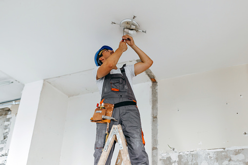 A young Caucasian male construction worker is standing on a ladder and using a screwdriver to fix a light bulb.