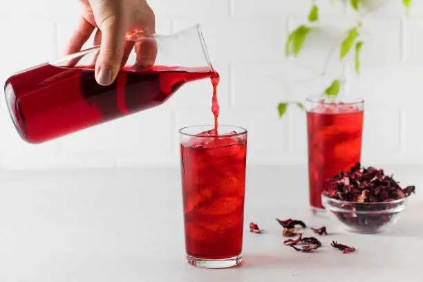 Photo of Herbal red tea made from hibiscus leaves is poured into a glass with ice from a jug.