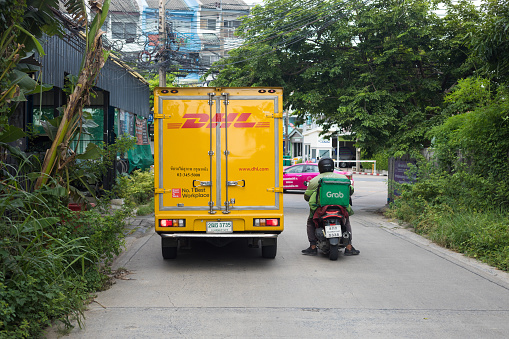 Thai express delivery motorcycle is overtaking a DHL car. Scene is in street in Bangkok Nawamin. Street is leading into a small intersection. At both sides are some plants and trees