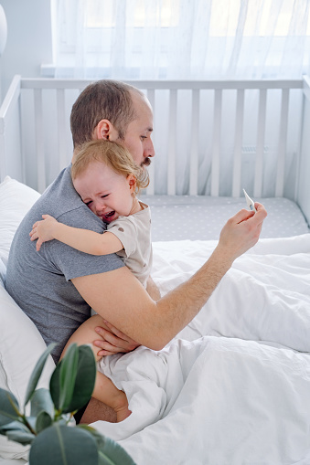 A young father holding his crying baby and checking temperature after measurement using thermometer.