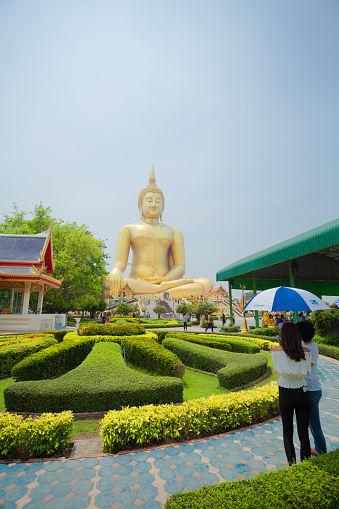 Giant golden thai buddha in park of temple grounds Wat Muang. In right foreground are some thai women with umbrella as sun protection. Temple is outside of  Ang Thong with national landmark 96m high Buddha statue of Thailand  in public free ground outside of city. In park are different themes of religion and places with statues