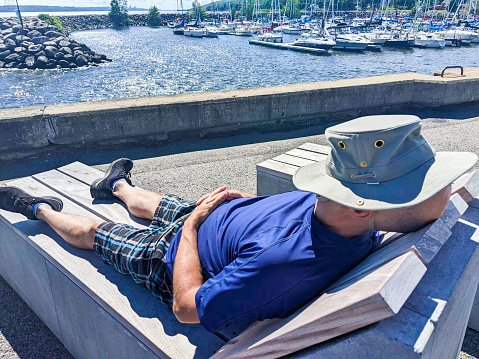 A man is sleeping on a wooden chaise longue with his hat on his face, on a wharf beside the St. Lawrence river in Quebec city during a nice day of summer