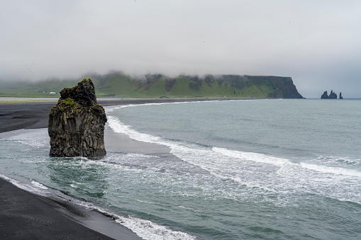 Reynisfjara Black Sand Beach on rainy, fogy day. Dramatic view on lonely clifs in the sea.