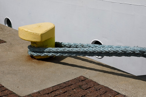Yellow-colored mooring bollard is located on the edge of the quay and is visible against the side of the moored ship. There is taut mooring rope on this bollard