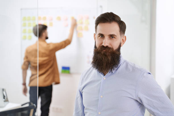 Happy smiling bearded senior developer or manager in modern IT office. Another professional working with scrum desk on background. Successful technology project or startup concept. High quality image stock photo