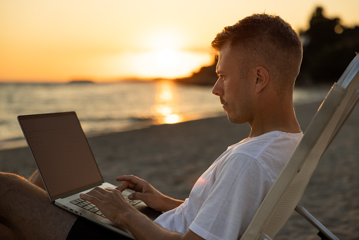 Caucasian young man working on laptop, on the beach during sunset, on his summer vacation