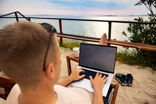 Caucasian young man working on laptop, on the beach during his summer vacation