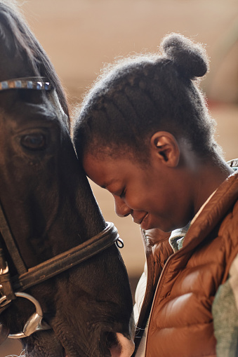 Vertical profile view of young black woman touching heads with beautiful horse in stables as bonding moment