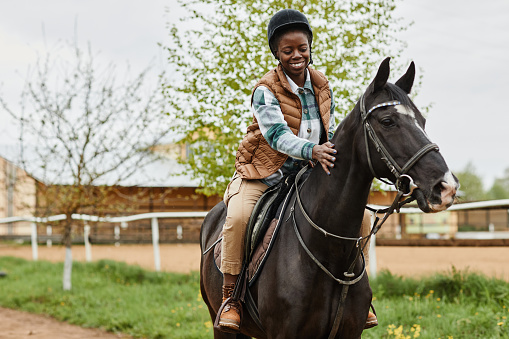 Portrait of smiling black woman riding horse in outdoor country ranch and stroking gently, copy space