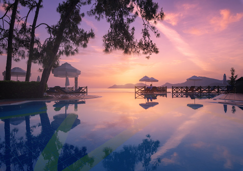 Beautiful reflection in swimming pool at colorful sunset. Purple sky reflected in water, palm trees, sun beds, umbrellas at night in summer. Luxury resort. Oludeniz, Turkey. Landscape with empty pool