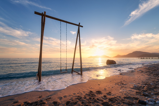Wooden swing in beautiful blue sea with waves, sandy beach, sky with golden sunlight at sunset in summer. Vacation in Oludeniz, Turkey. Tropical landscape with swing on sea coast, water. Travel