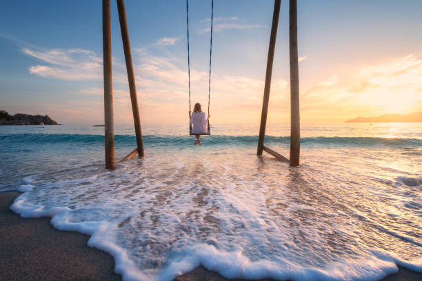 Happy young woman on wooden swing in water, beautiful blue sea with waves, sandy beach, golden sky at sunset. Summer holiday in Oludeniz, Turkey. Girl ride on a swing on sea coast, clear water. Travel stock photo