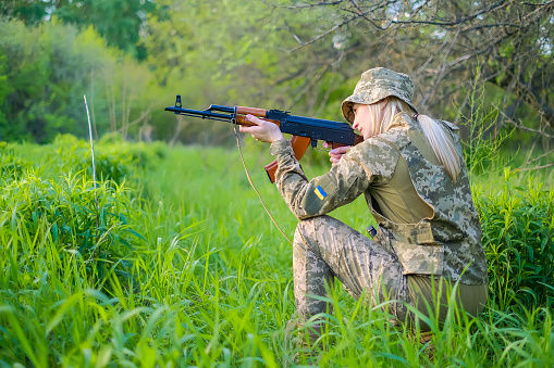 Young Ukrainian female soldier in camouflage uniform with an assault rifle in reconnaissance patrols a combat zone and takes aim while kneeling. Brave military patriot woman with a gun in the forest