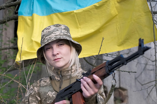 Armed with assault rifle determined female soldier in camouflage uniform looking at the camera on the waving Ukrainian flag background. Ukrainian woman patriot.