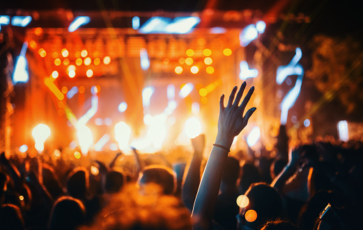 Rear view of large group of people enjoying a concert performance. There are many hands applauding and taping the show. Multi colored lasers and spot lights firing from the stage.\nSilhouettes have been significantly liquified.