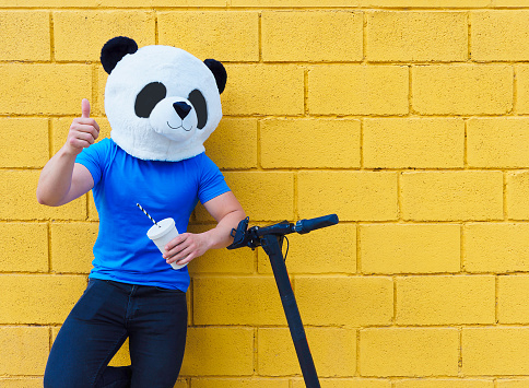 Man with panda mask making ok gesture leaning on electric scooter with drink in other hand. Yellow wall background.