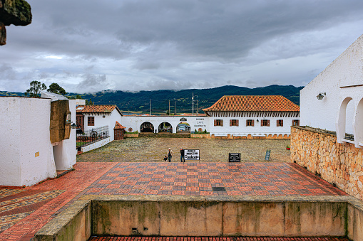 Colombia, South America - July 08, 2022: View of the main town square in the Andes town of Guatavita in the Cundinamarca Department of the South American country of Colombia, from the entrance to the square. Colonial Spanish style of architecture with terracota tiled roof is seen around the square. In the far background are the Tominé reservoir and the Andes Mountains. The altitude is 8,790 feet above mean sea level. Photo shot in the afternoon sunlight on an overcast day;  horizontal format. Copy space.