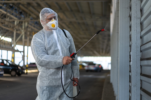 Worker wearing protective suit disinfection gear disinfect surface public place parking. Side view of disinfector safety overall and respiratory mask Covid pandemic disinfection preventive measures