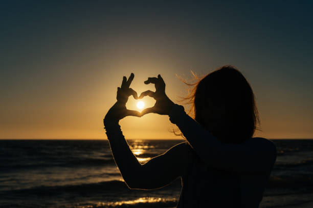 istock woman making a heart with her fingers against the light while the sun is setting 1408537311