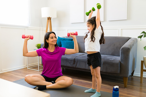 Excited fit mother working out at home while lifting dumbbell weights with her adorable little kid