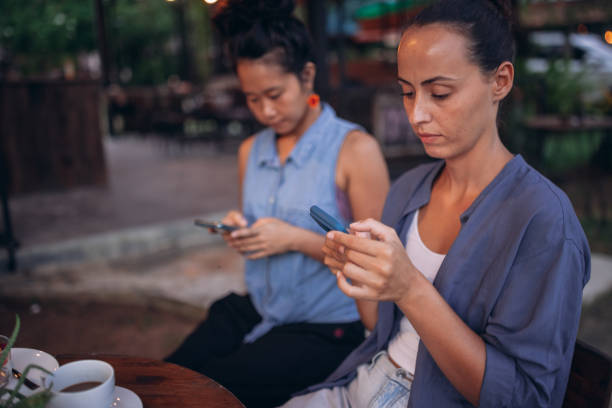 Social media changes the way you hang out with friends Female friends are sitting in a cafe and using their phones, ignoring each other. wasting time stock pictures, royalty-free photos & images