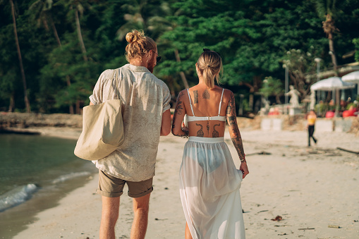 Rear view of a smiling couple is walking on a beach at sunset.