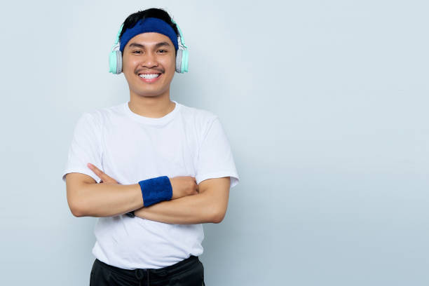 улыбающийся молодой азиатский спортсмен в синей повязке на голове и белой футболке спортивной одежды во время прослушивания любимой музык - arms crossed audio стоковые фото и изображения